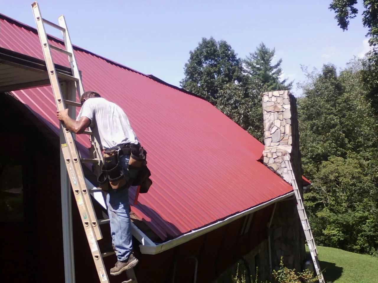 Roofer on a ladder beside a red metal roof and stone chimney — Soffit & Fascia Repair by Roofing Brothers in Burden